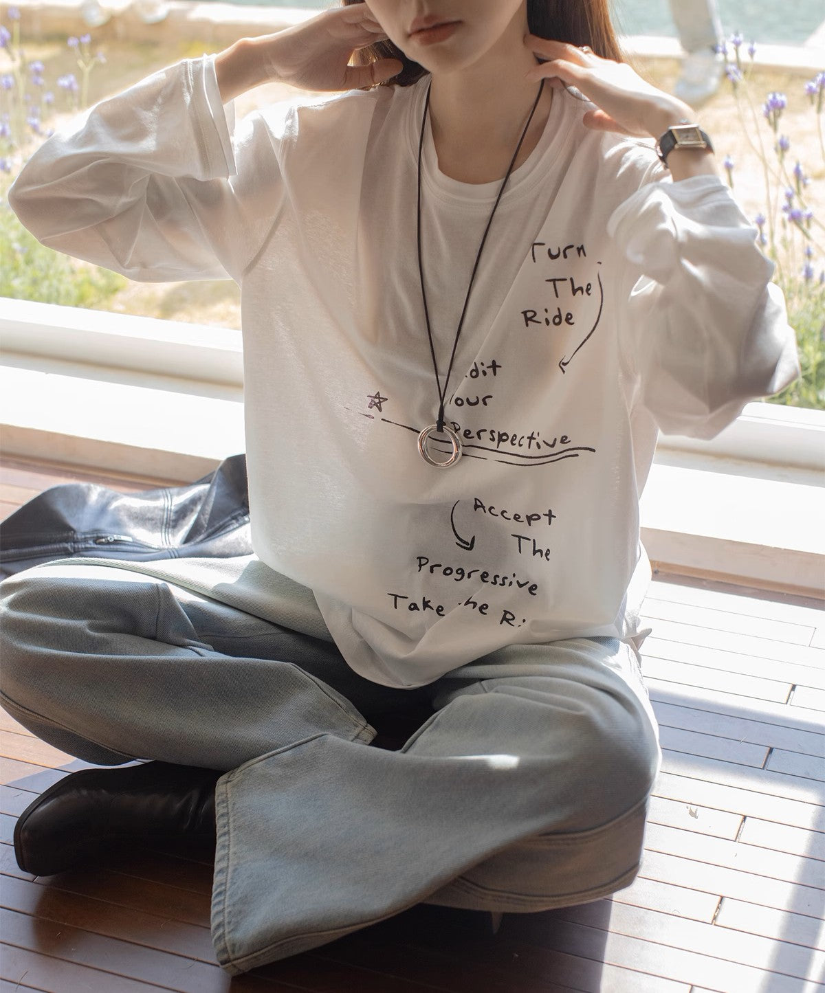 白のメッセージプリントロングTシャツとワイドパンツを着用し、窓辺に座っている女性。オーバーサイズ感が特徴的。