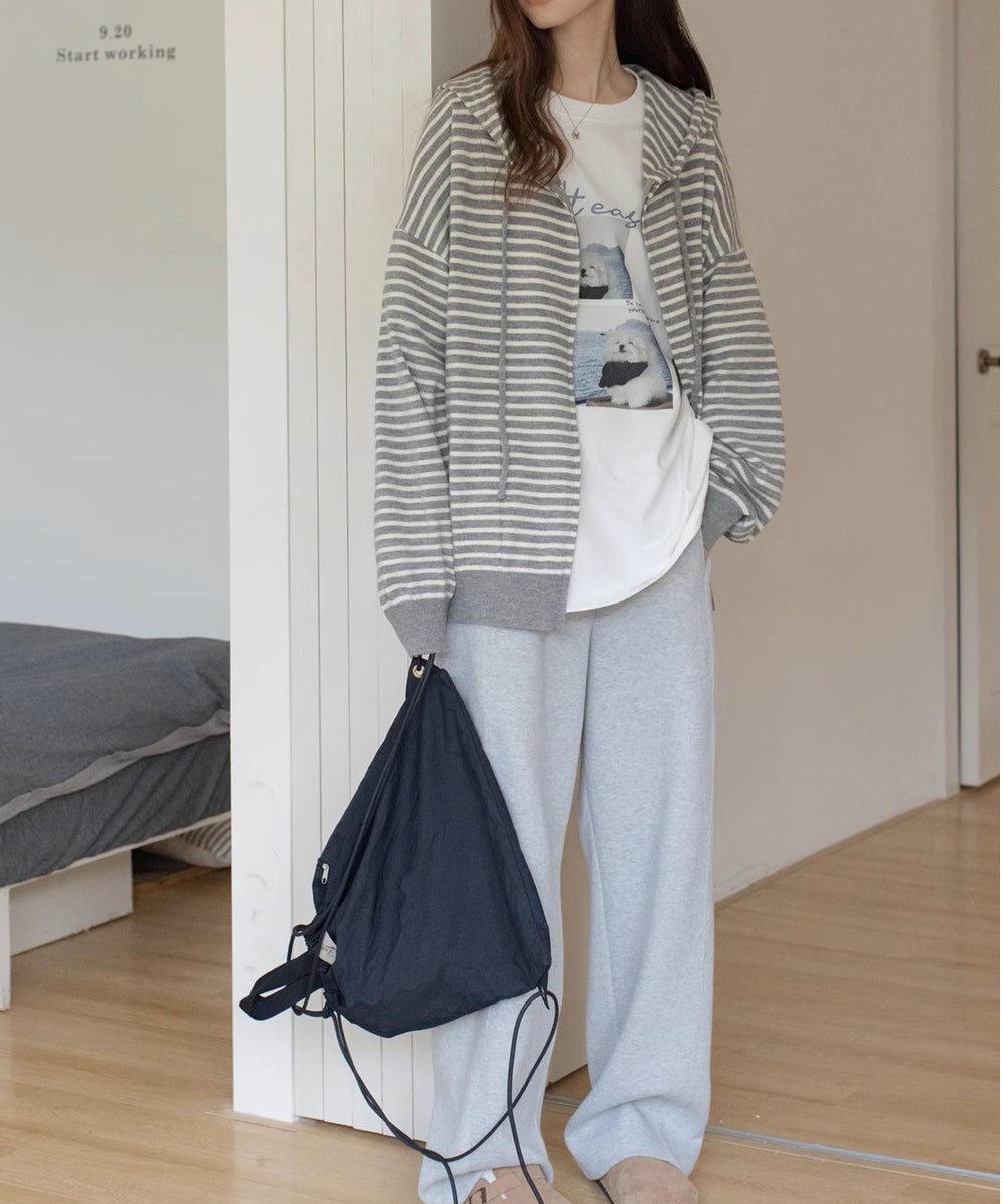 グレーと白の細いボーダー柄のジップアップパーカーを着用した女性の全身。インナーには白いTシャツ、ボトムスにはライトグレーのスウェットパンツを合わせたカジュアルな着こなし。黒いバッグを持っている。