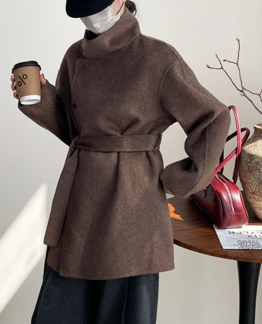 ブラウンのコートを着てカフェのテーブル横に立つ女性。襟を立てたデザインと横顔のシルエット、暖かみのある素材感。