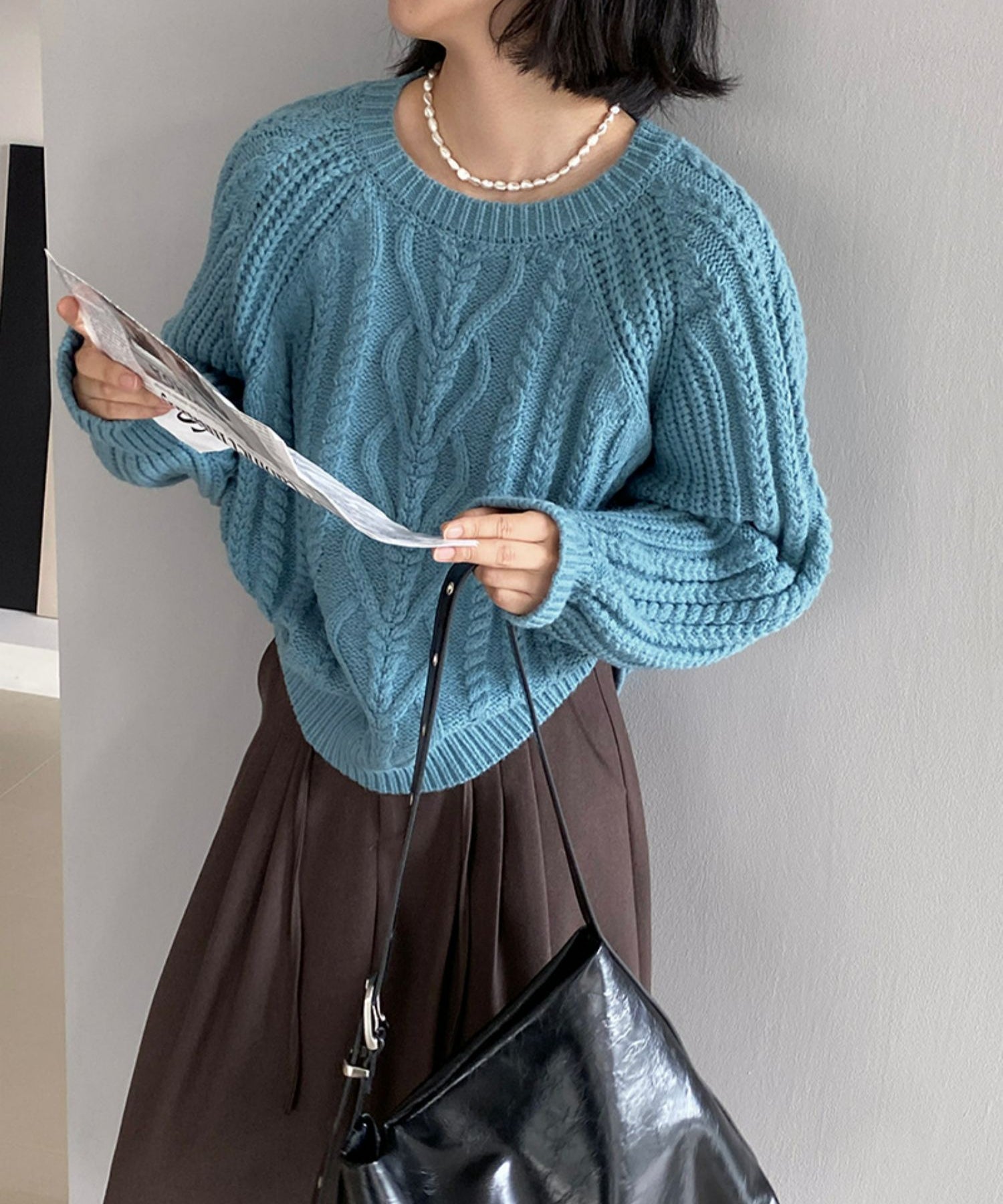 ブルーのケーブルニットを着用し、茶色のプリーツスカートとパールのネックレスを合わせた女性。黒いバッグをさげている。