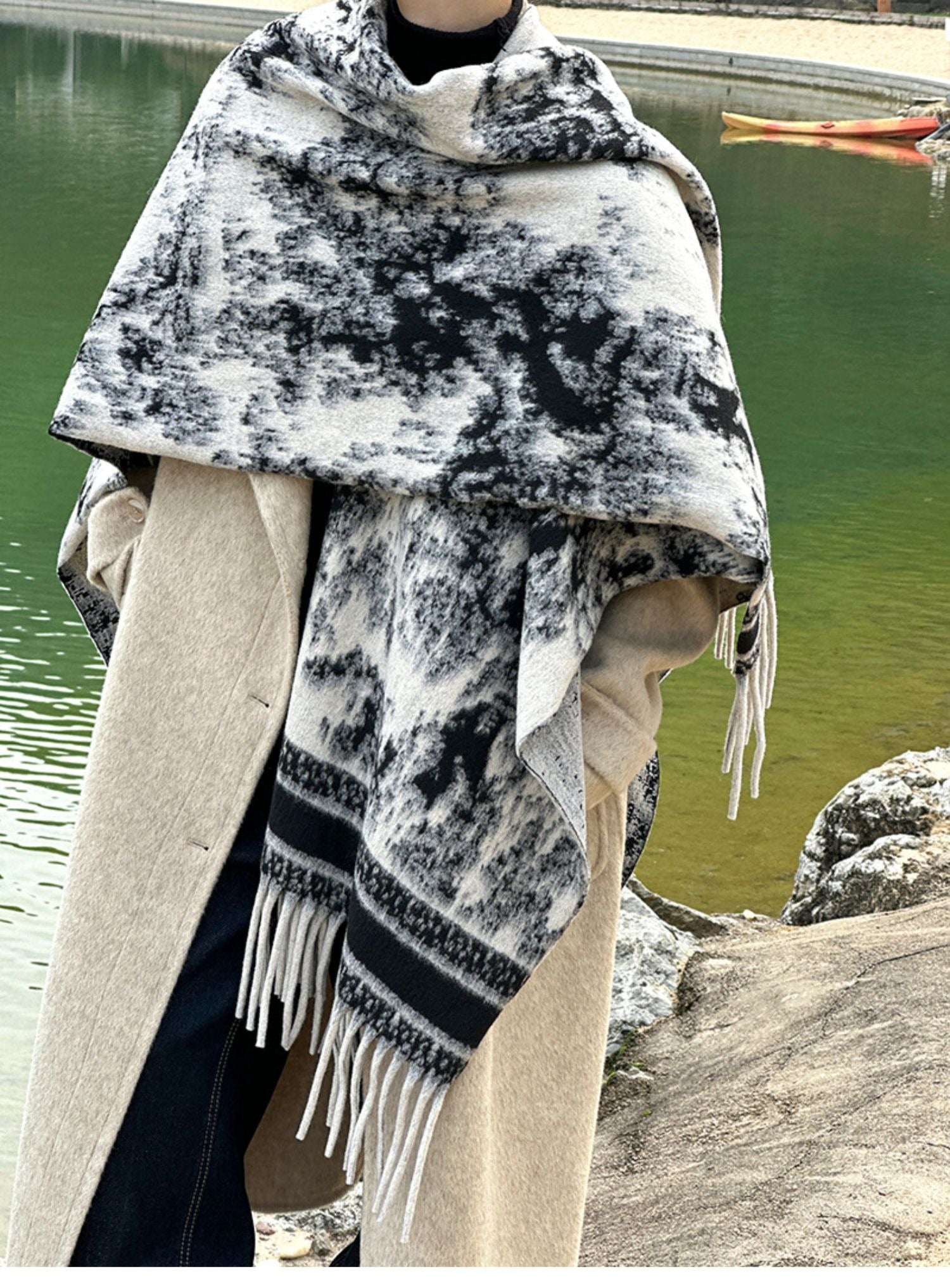 モノトーンの水墨画風の柄ショールをベージュのコートと合わせて屋外で着用する女性が池の前に立つ写真
