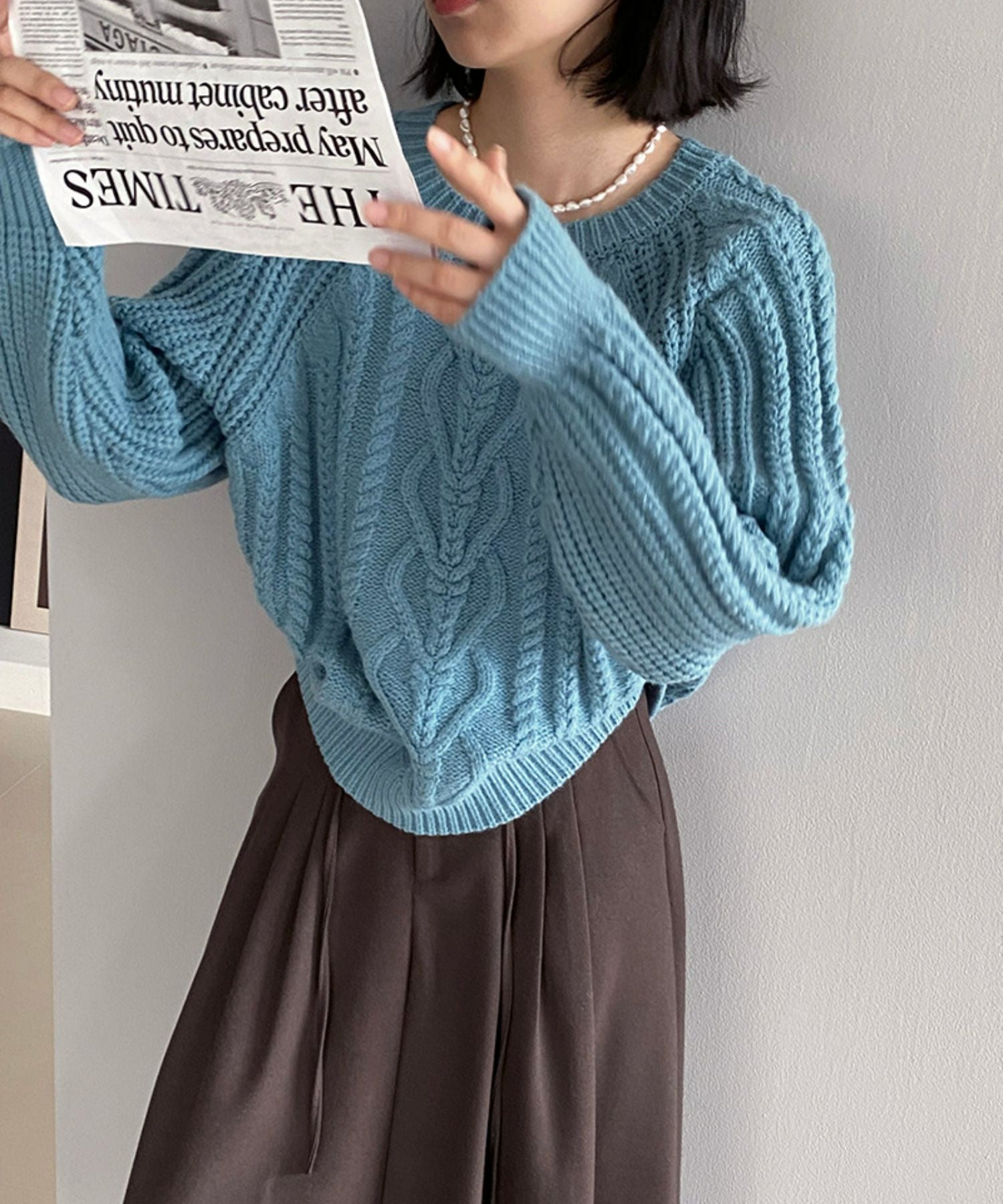 ブルーのケーブルニットを着用し、茶色のプリーツスカートとパールのネックレスを合わせた女性。英字の新聞を見ている。7