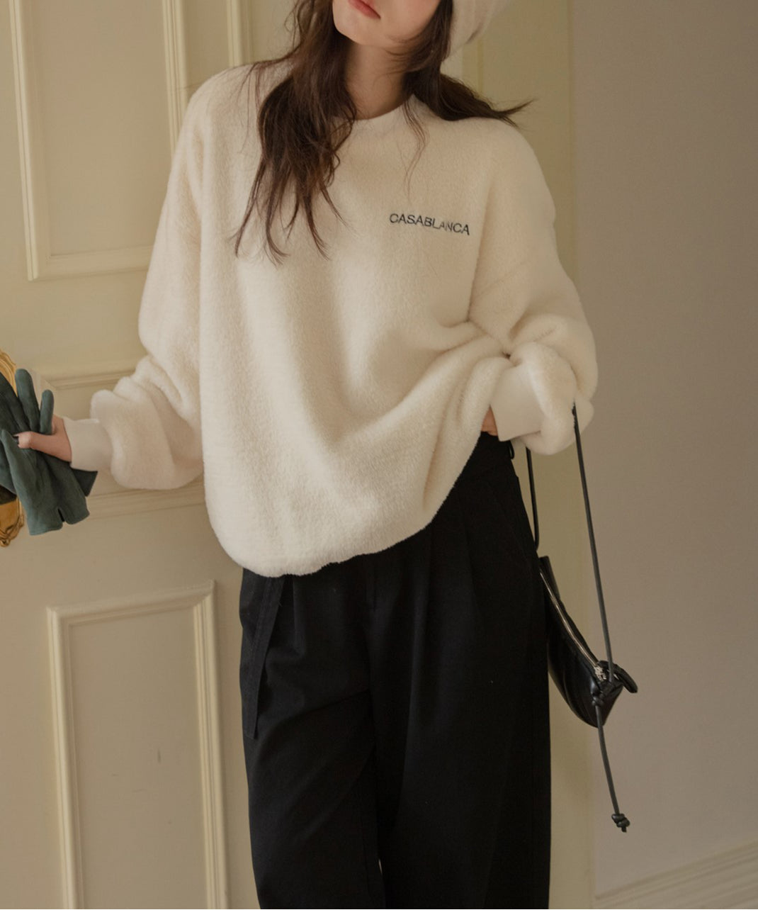 オフホワイトのふわふわしたボア素材のプルオーバーを着た女性のクローズアップ。手に手袋を持っている。左胸には「CASABLANCA」の刺繍ロゴが見える。