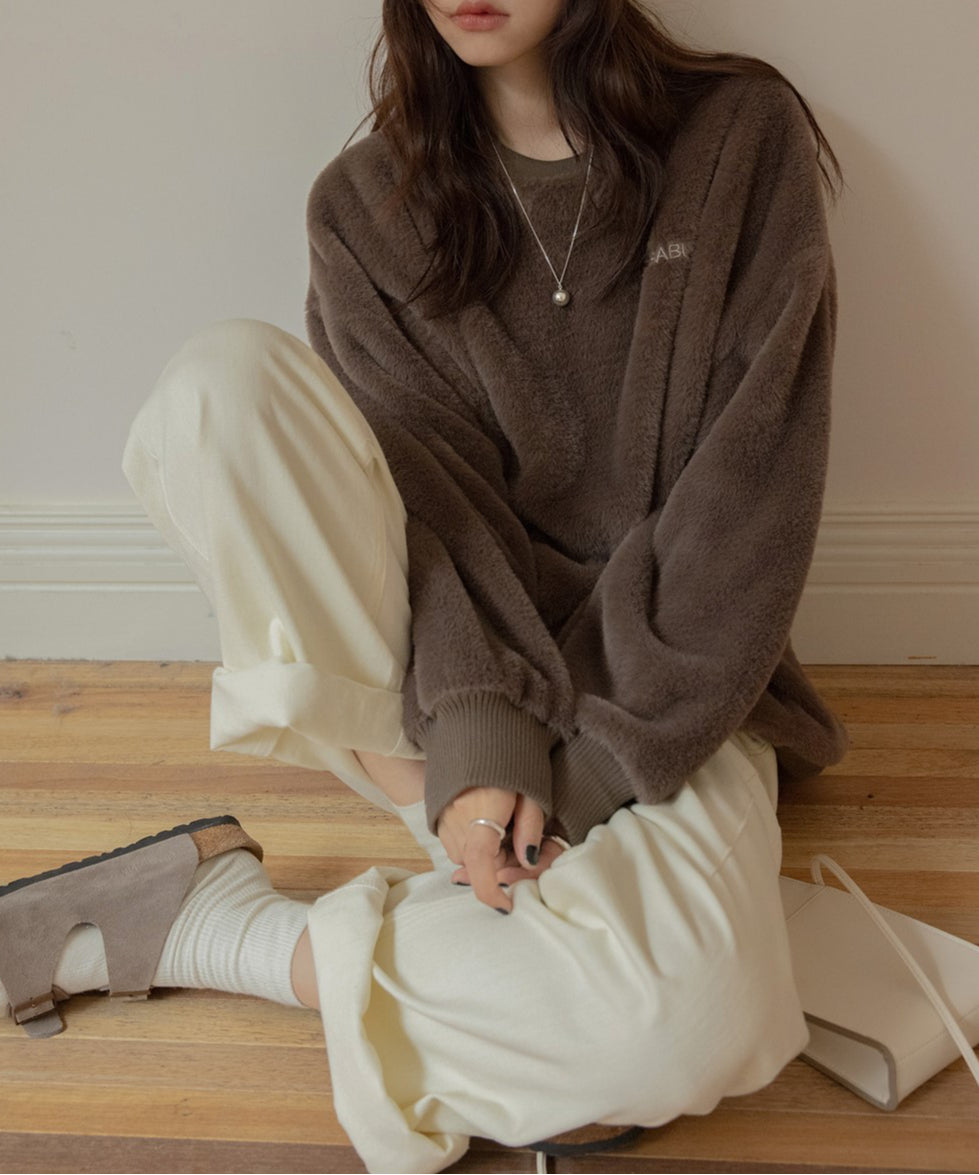 ブラウン色のふわふわしたボア素材のプルオーバーを着た女性が室内に座っている。胸の刺繍ロゴが見える。