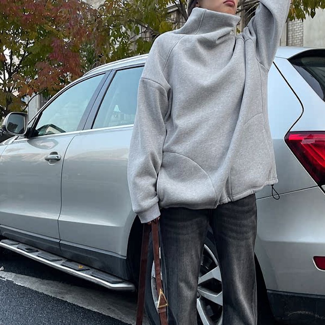 グレーのタートルネックスウェットと濃いグレーのデニムを着用した人物の全身写真。シルバーのSUV車の横に立っている。