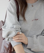 グレーのオーバーサイズスウェットを着用した女性のクローズアップ。胸元には「Summer Night」の赤茶色の刺繍ロゴがあり、左手にシルバーのリングとバングルを着用し、茶色のバッグを肩にかけている。