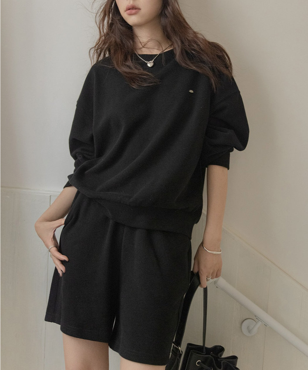 黒のセットアップを着用した女性の半身。手には黒いバッグを持っている。