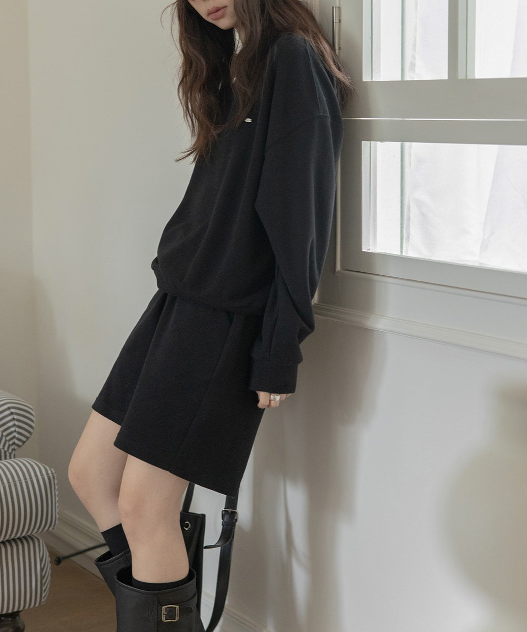 黒のセットアップを着用した女性。黒のハイソックスとロングブーツ、シルバーのネックレスを着用している。