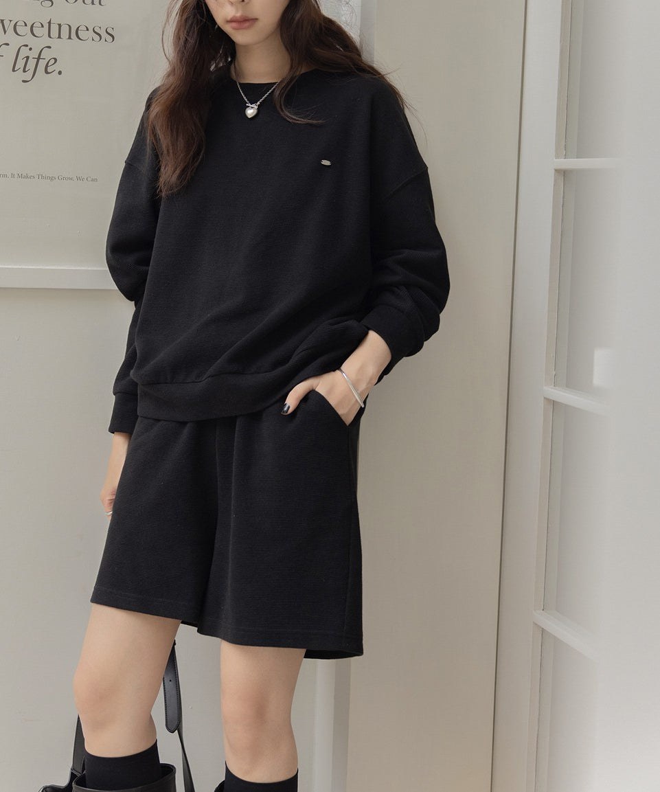 黒のセットアップを着用し、片手をポケットに入れた女性の全身。黒のハイソックスとロングブーツ、シルバーのバングル、ネックレスを着用している。