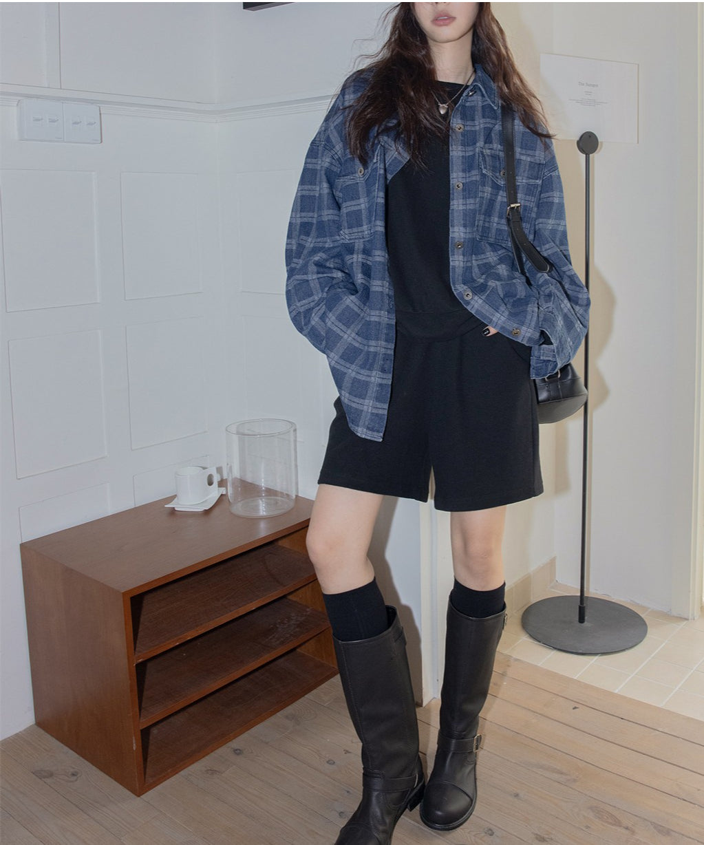 青と黒のチェック柄オーバーサイズシャツを羽織り、黒のTシャツ、黒のショートパンツ、黒のハイソックス、黒のロングブーツを着用した女性の全身コーディネート。黒のショルダーバッグを持ち、室内の壁際で立っている。
