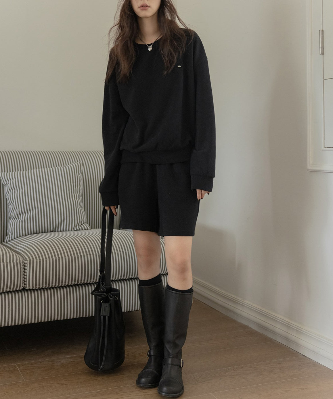 黒のセットアップを着用した女性の全身。黒のハイソックスとロングブーツ、ネックレスを着用している。