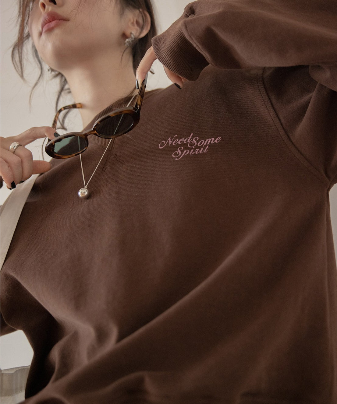 ブラウンのスウェットを着用した女性のクローズアップ。胸元にピンク色の刺繍ロゴがあり、ネックレスとサングラスを持っている。