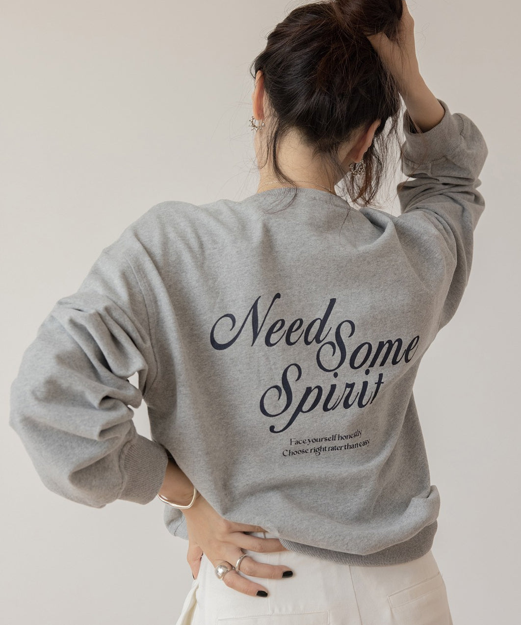 グレーのスウェットを着用した女性の背部のクローズアップ。ネイビーの刺繍ロゴがあり、白いワイドパンツを履いている。