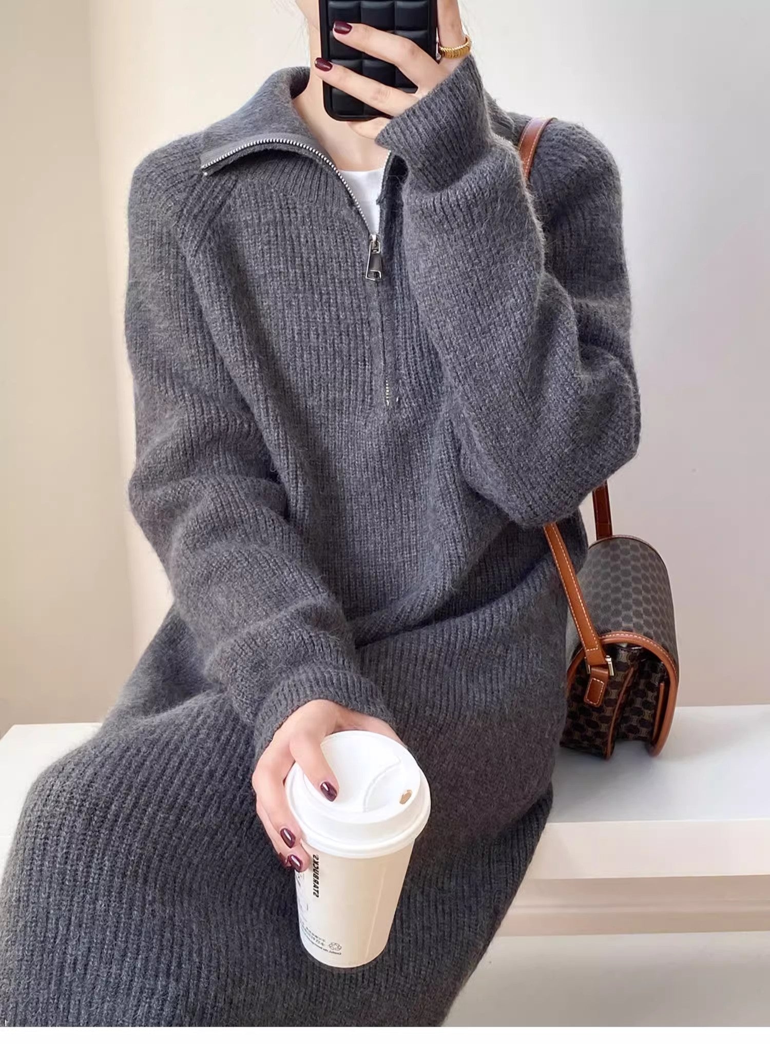 グレーのハーフジップニットロングワンピースを着用し、茶色のバッグと白いコーヒーカップを持つ女性のクローズアップ画像。