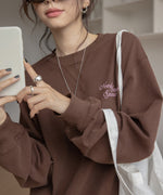 ブラウンのスウェットを着用した女性のクローズアップ。胸元にピンク色の刺繍ロゴがあり、スマホを手に持っている。
