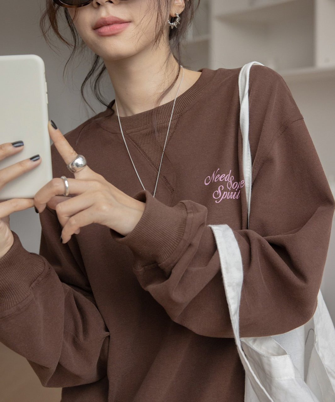 ブラウンのスウェットを着用した女性のクローズアップ。胸元にピンク色の刺繍ロゴがあり、スマホを手に持っている。