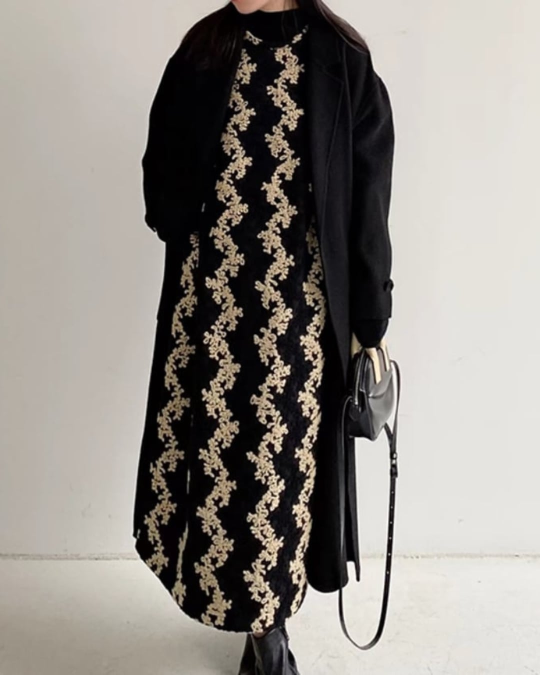 黒いロングコートの下に花柄のジャンパースカートと黒いタートルネックを着用した女性の全体画像。